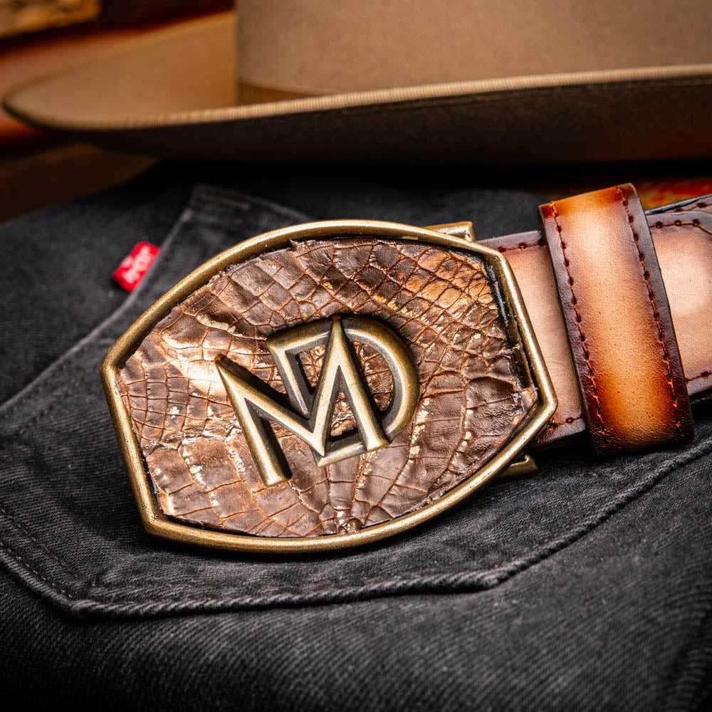 The Texas Belt Stone Brown by Marco Di Milano features embossed brown leather and a large gold-and-brown "CW" buckle, styled after a classic western calfskin belt with "MARCO DI MILANO" branding on the strap.