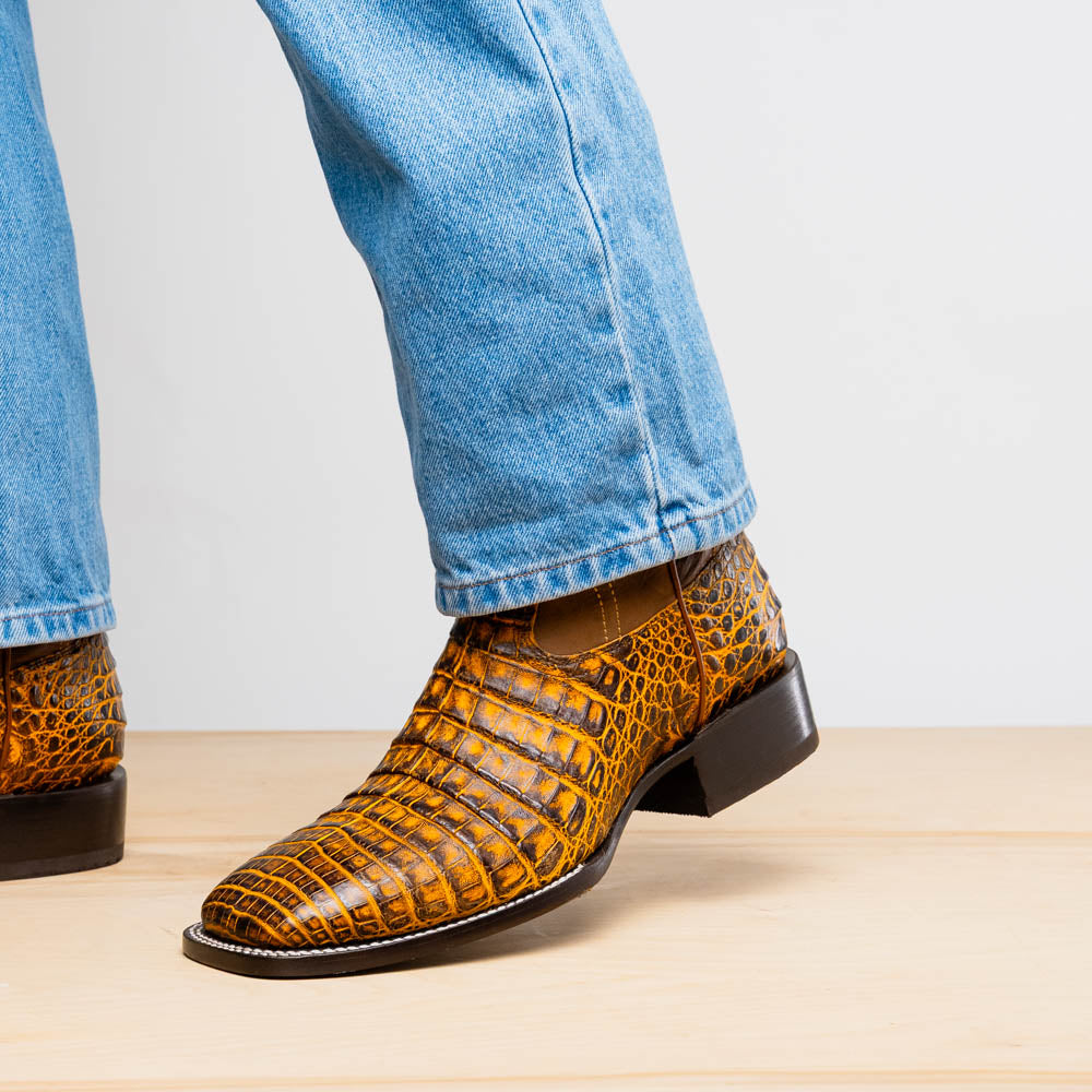 The Marco Di Milano Tucson Mustard Brown men's Western boots feature brown leather uppers and yellow genuine caiman belly skin lowers; one boot stands upright, the other shows the signature square toe cowboy boot sole.