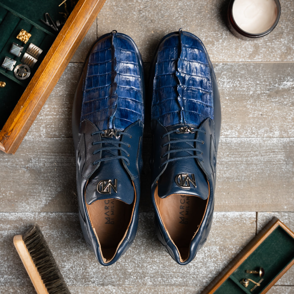 Introducing the Udine Navy by Marco Di Milano—a pair of luxury men’s sneakers crafted from exotic navy blue leather with textured uppers and durable rubber soles. One shoe stands upright while the other rests on its side, showcasing their distinctive style and impeccable craftsmanship.