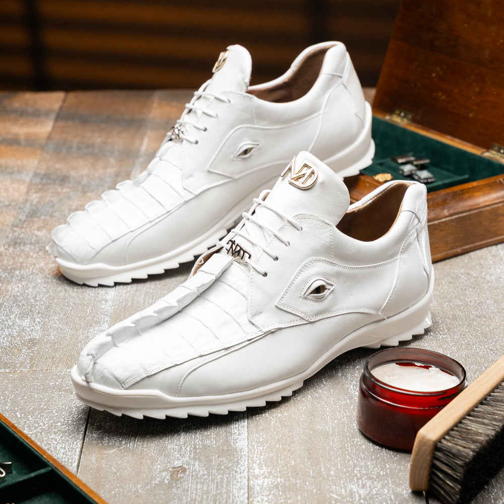 The Udine White by Marco Di Milano is a pair of luxury men’s sneakers crafted from white hornback caiman leather with a distinctive crocodile pattern, featuring a gold logo on the tongue and textured rubber soles. Displayed with one shoe upright and the other leaning against it, this footwear exudes sophistication and unique style.