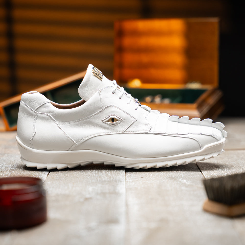 The Udine White by Marco Di Milano is a pair of luxury men’s sneakers crafted from white hornback caiman leather with a distinctive crocodile pattern, featuring a gold logo on the tongue and textured rubber soles. Displayed with one shoe upright and the other leaning against it, this footwear exudes sophistication and unique style.