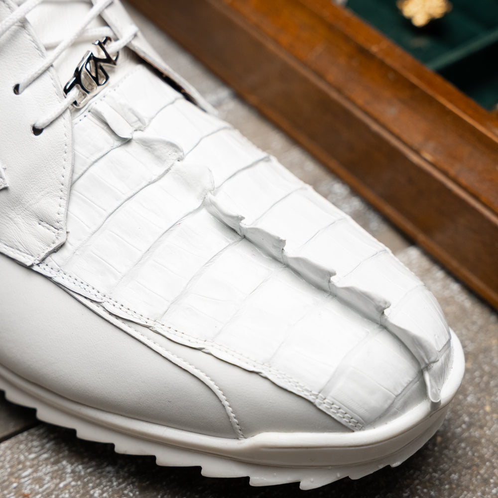 The Udine White by Marco Di Milano is a pair of luxury men’s sneakers crafted from white hornback caiman leather with a distinctive crocodile pattern, featuring a gold logo on the tongue and textured rubber soles. Displayed with one shoe upright and the other leaning against it, this footwear exudes sophistication and unique style.