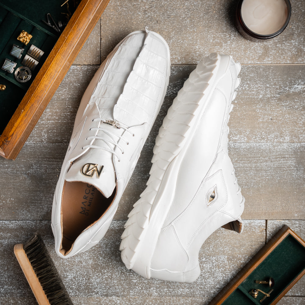 The Udine White by Marco Di Milano is a pair of luxury men’s sneakers crafted from white hornback caiman leather with a distinctive crocodile pattern, featuring a gold logo on the tongue and textured rubber soles. Displayed with one shoe upright and the other leaning against it, this footwear exudes sophistication and unique style.
