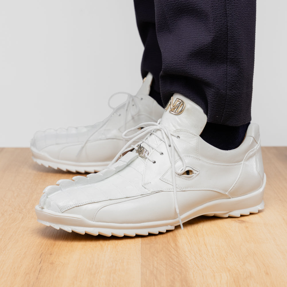 The Udine White by Marco Di Milano is a pair of luxury men’s sneakers crafted from white hornback caiman leather with a distinctive crocodile pattern, featuring a gold logo on the tongue and textured rubber soles. Displayed with one shoe upright and the other leaning against it, this footwear exudes sophistication and unique style.