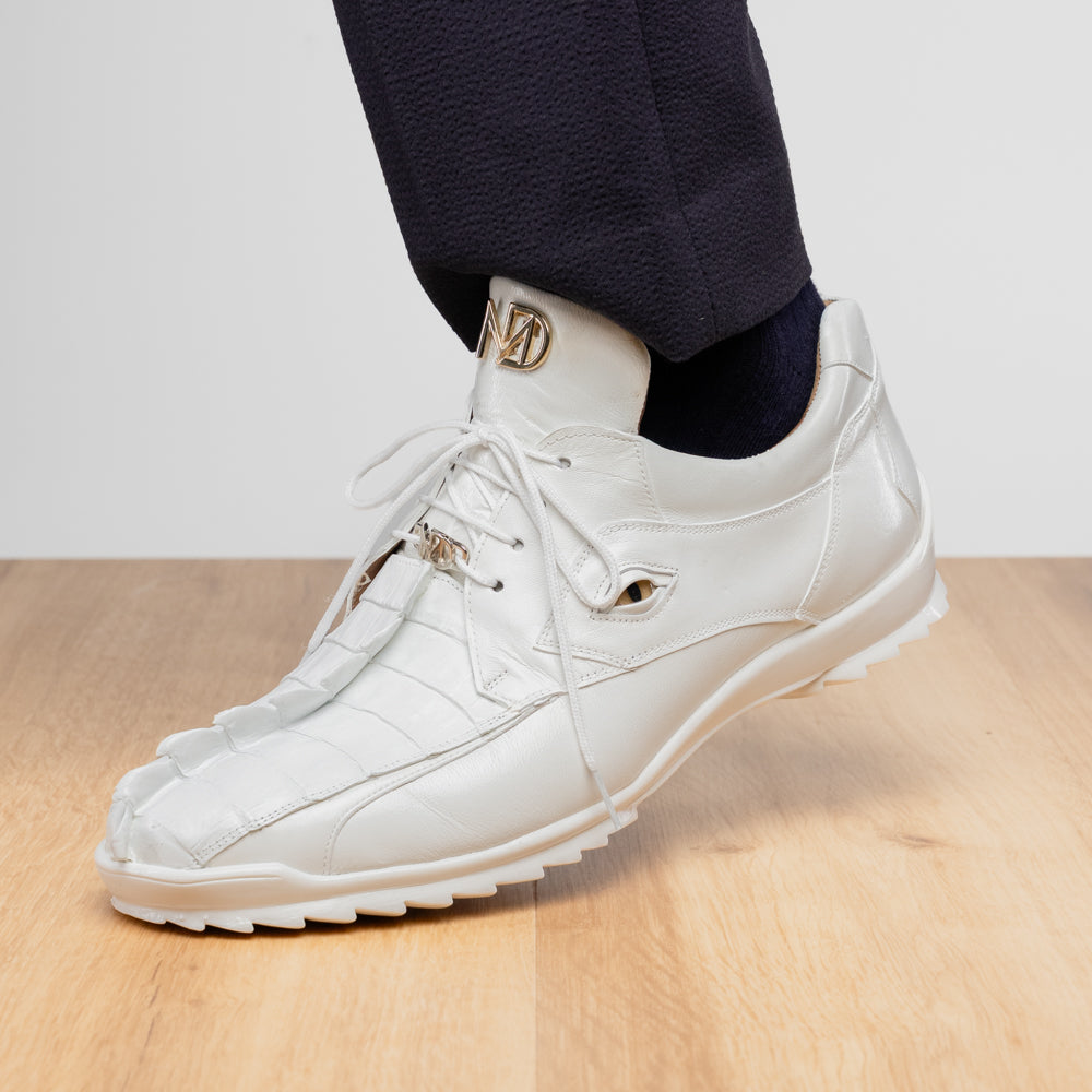 The Udine White by Marco Di Milano is a pair of luxury men’s sneakers crafted from white hornback caiman leather with a distinctive crocodile pattern, featuring a gold logo on the tongue and textured rubber soles. Displayed with one shoe upright and the other leaning against it, this footwear exudes sophistication and unique style.