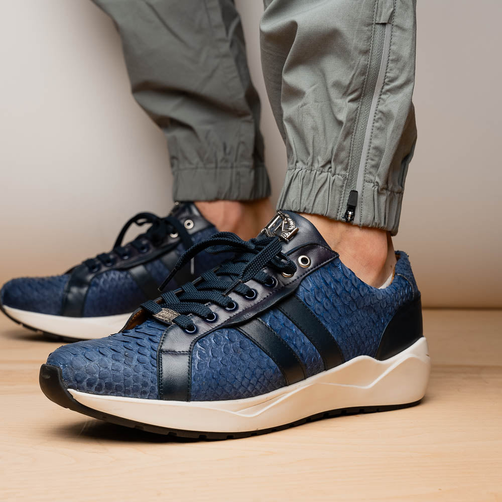 The Verona Navy sneakers by Marco Di Milano feature a blue python texture, navy laces, side stripes, and white rubber soles—luxury footwear with one shoe upright and the other on its side.