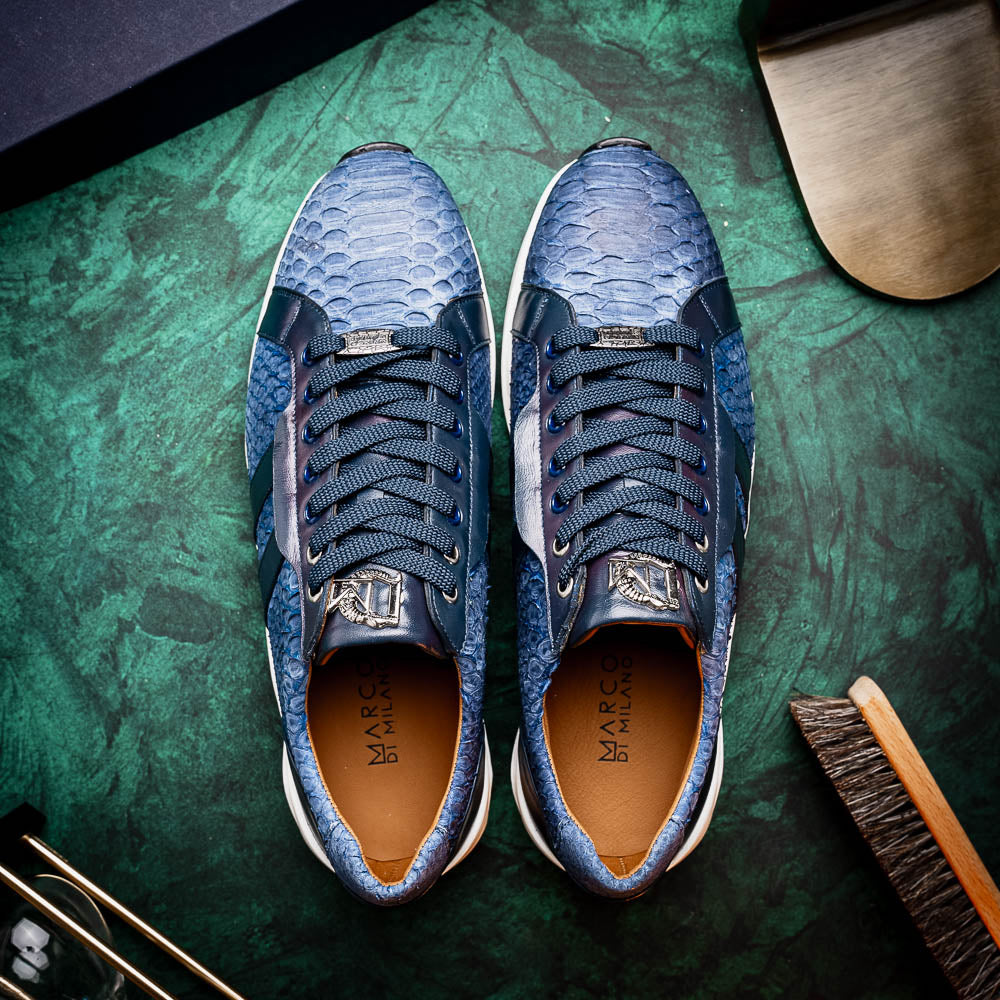 The Verona Navy sneakers by Marco Di Milano feature a blue python texture, navy laces, side stripes, and white rubber soles—luxury footwear with one shoe upright and the other on its side.