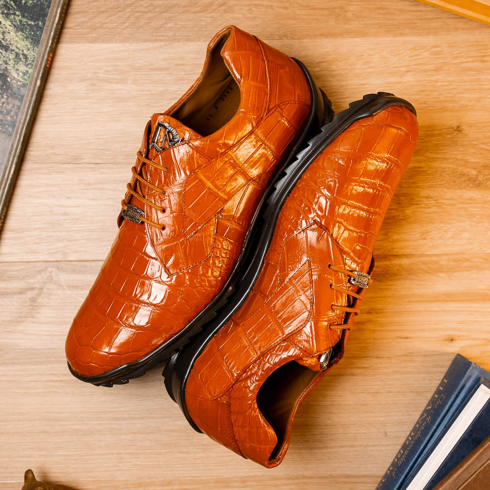 The Marco Di Milano Vulcan Honey features orange, crocodile-patterned leather dress shoes with hand-painted finishes and black rubber soles; one shoe stands upright while the other rests on its side below.