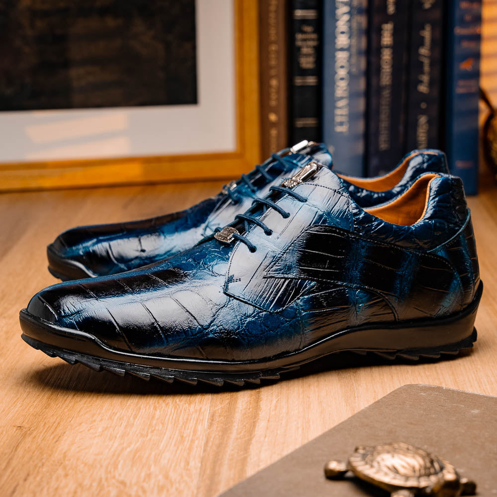 The Vulcan Rustic Navy by Marco Di Milano features glossy blue and black lace-up dress shoes with textured leather, rubber soles, and exquisite hand-painted finishes—one upright, the other propped beneath it—showcasing luxury footwear.