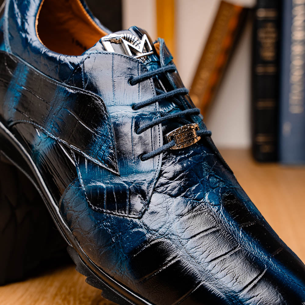 The Vulcan Rustic Navy by Marco Di Milano features glossy blue and black lace-up dress shoes with textured leather, rubber soles, and exquisite hand-painted finishes—one upright, the other propped beneath it—showcasing luxury footwear.