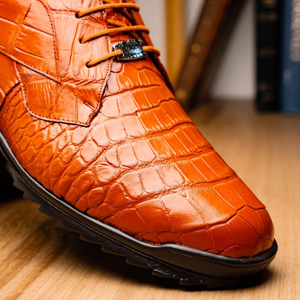 The Marco Di Milano Vulcan Honey features orange, crocodile-patterned leather dress shoes with hand-painted finishes and black rubber soles; one shoe stands upright while the other rests on its side below.
