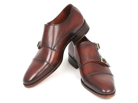 The Paul Parkman Captoe Double Monkstraps Brown (Model 0457-BRW) are handcrafted from exquisite calfskin leather, featuring two elegant silver buckles on each shoe. One shoe leans casually against the other, highlighting their timeless style.