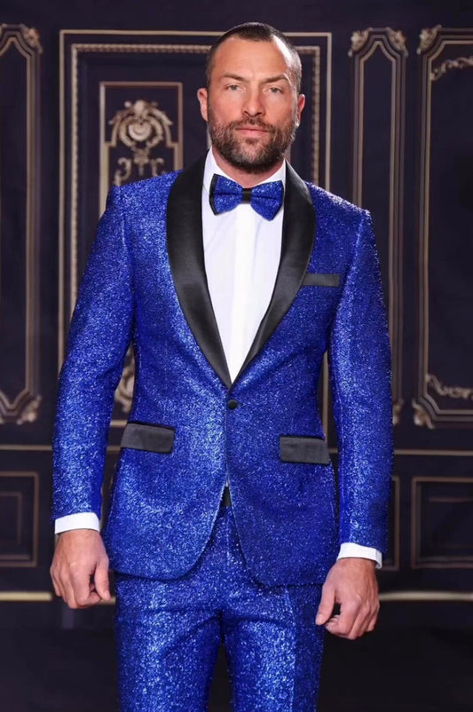 A man in a Manzini 2-piece slim fit suit featuring a satin shawl collar in royal blue stands against a decorative backdrop, complemented by a matching bow tie.