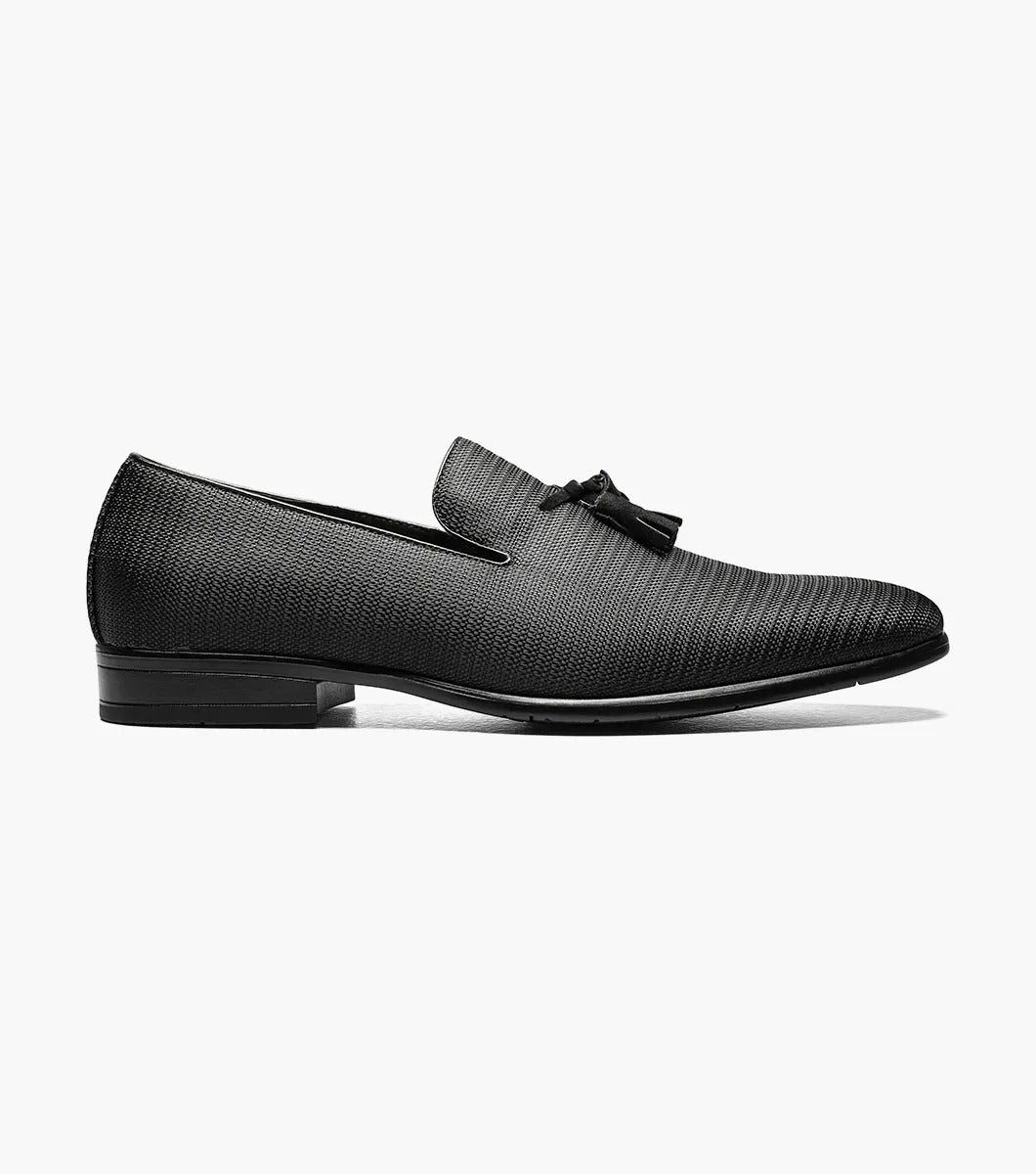 The STACY ADAMS Tazewell Plain Toe Tassel Slip On in black (25343-001) features textured leather, decorative tassels, a low heel, and cushioned footbed, shown on a white background.