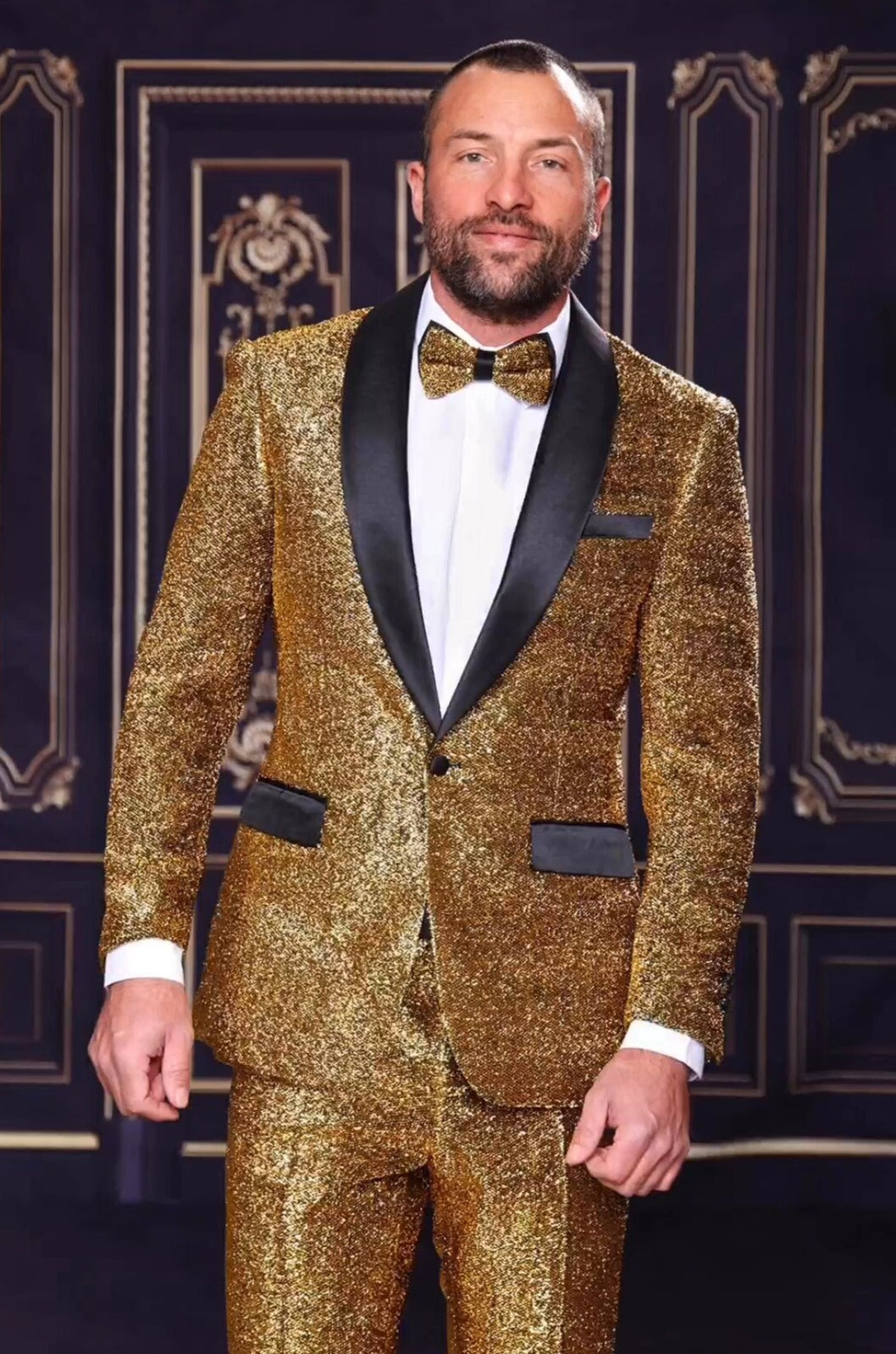 A person dressed in a Manzini 2pc Slim Fit Gold Suit with a satin shawl collar, complemented by a crisp white shirt and sleek bow tie, stands against a dark, decorative background.