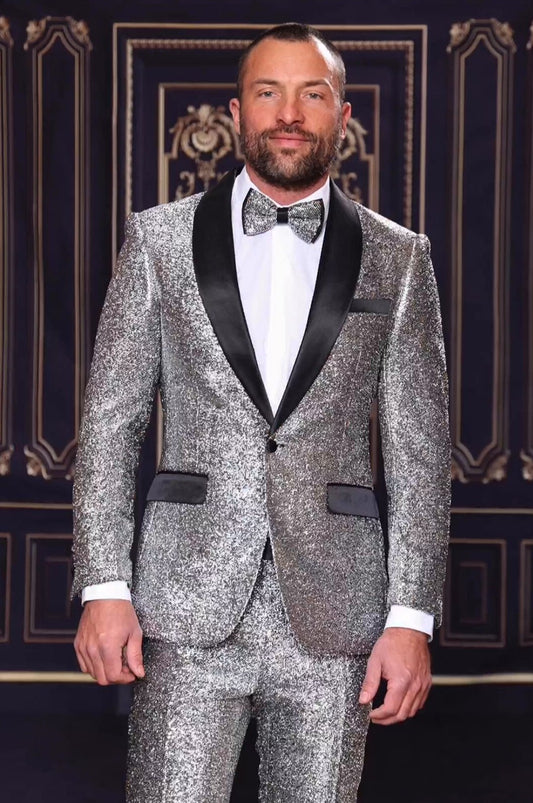 A man in a MANZINI 2pc Slim Fit with Satin Shawl Collar Silver Suit gracefully complements his bow tie, standing against a decorative dark background.