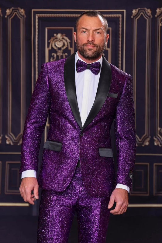 A man dressed in a Manzini 2pc Slim Fit Purple Suit, featuring a satin shawl collar, and accessorized with a striking bow tie, stands confidently against an ornate background.