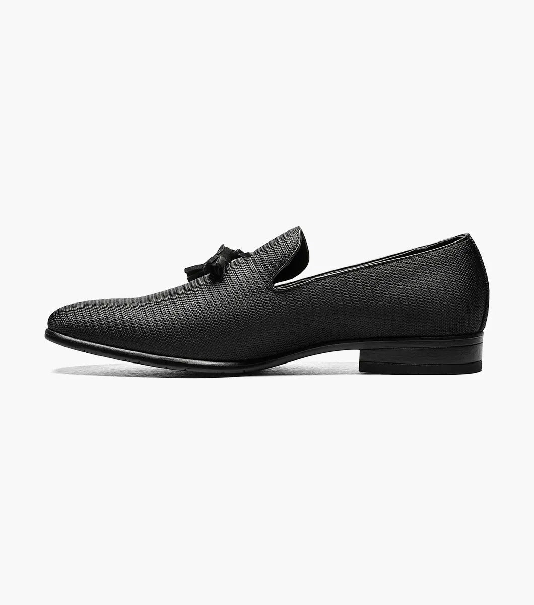 The STACY ADAMS Tazewell Plain Toe Tassel Slip On in black (25343-001) features textured leather, decorative tassels, a low heel, and cushioned footbed, shown on a white background.