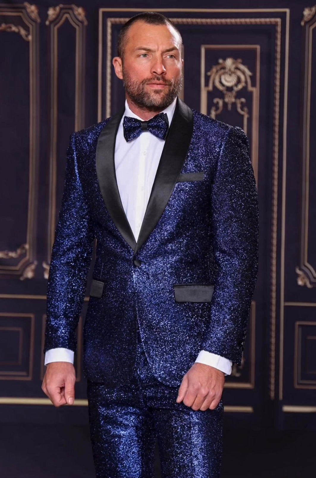 A man wearing the Manzini 2pc Slim Fit with Satin Shawl Collar Midnight Suit stands against an ornate backdrop, exuding timeless elegance. His sophisticated look is perfect for winter soirées and can effortlessly transition into a sleek summer suit style with just minor accents.