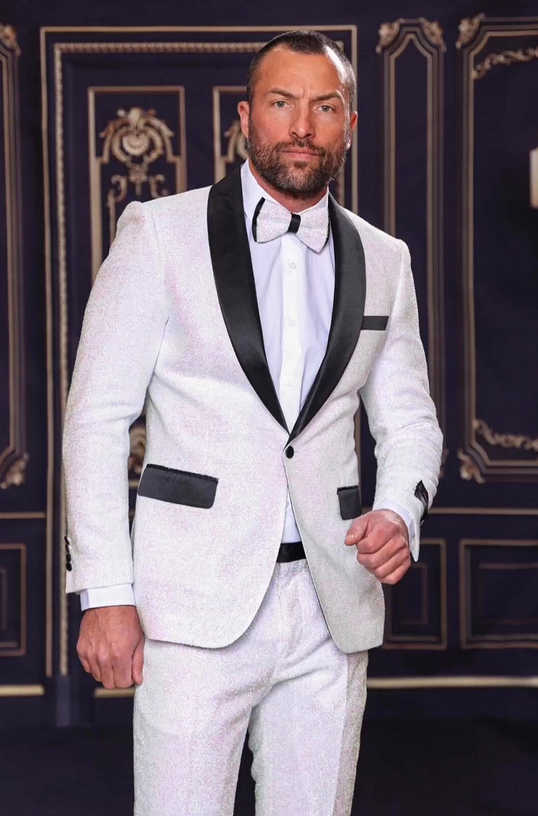 A gentleman wearing the MANZINI 2pc Slim Fit with Satin Shawl Collar White Suit from Manzini stands before an ornate dark background, complemented by a classic bow tie.