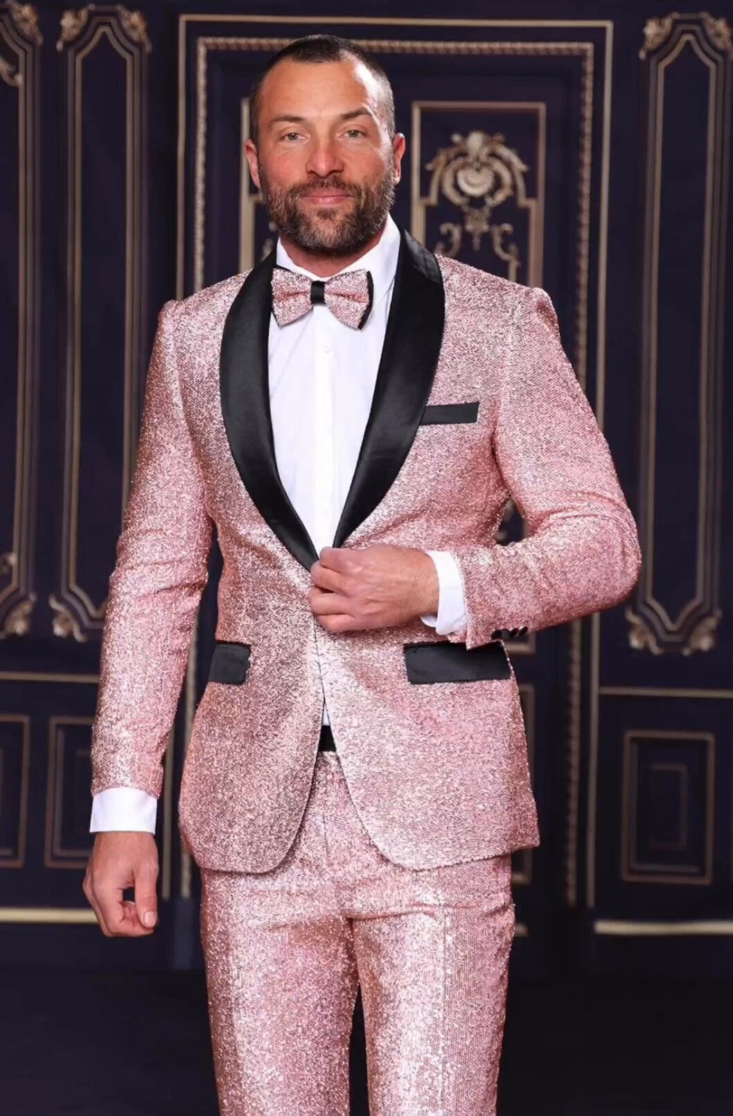 A bearded man in a MANZINI 2pc Slim Fit with Satin Shawl Collar Rose Gold Suit by Manzini, complemented by a matching bow tie, stands against a dark, ornate background.