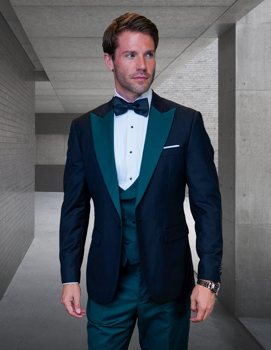 A man in a formal tuxedo suit from Statement Clothing's STATEMENT CLOTHING | ARYA-HUNTER collection, featuring an Italian fabric black jacket, green vest, and bow tie, stands in a modern corridor.