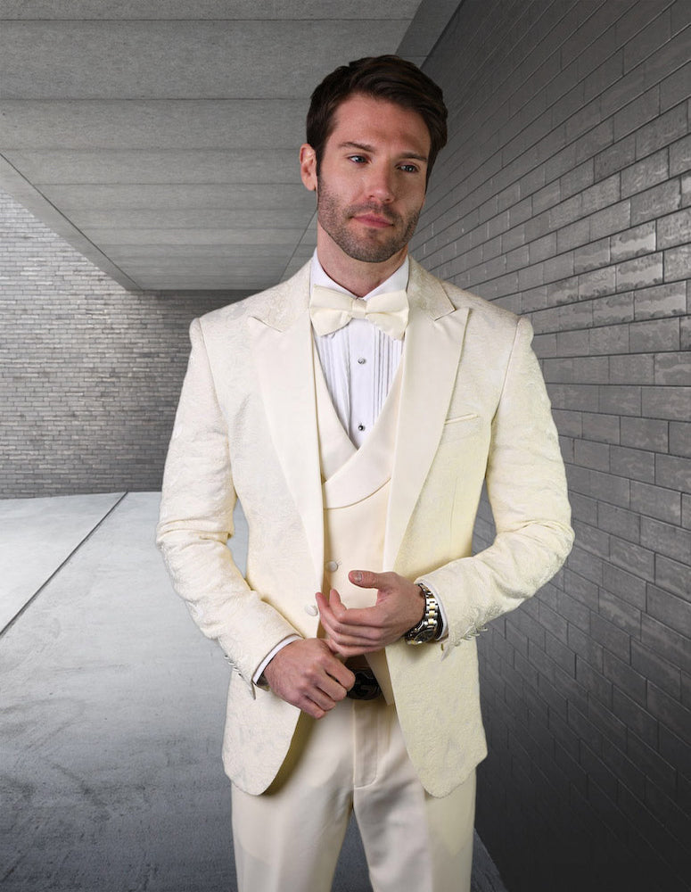A man wearing the Belagio15-Offwht tuxedo from Statement Clothing, complete with a bow tie and flat front pants, stands in a corridor lined with gray brick walls.