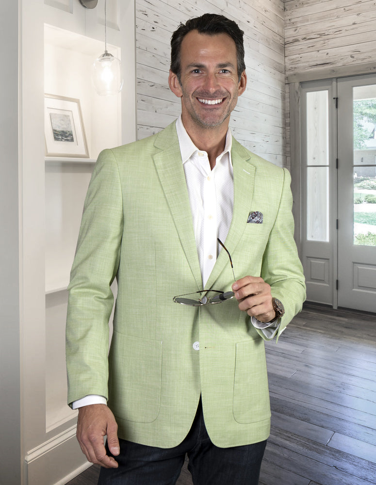 A man in the BELUCHI-MINT suit jacket from Statement Clothing, crafted with SUPER 150'S ITALIAN FABRIC, holds glasses while standing in a modern, well-lit room with wooden flooring and a BELUCHI picture frame on the wall.