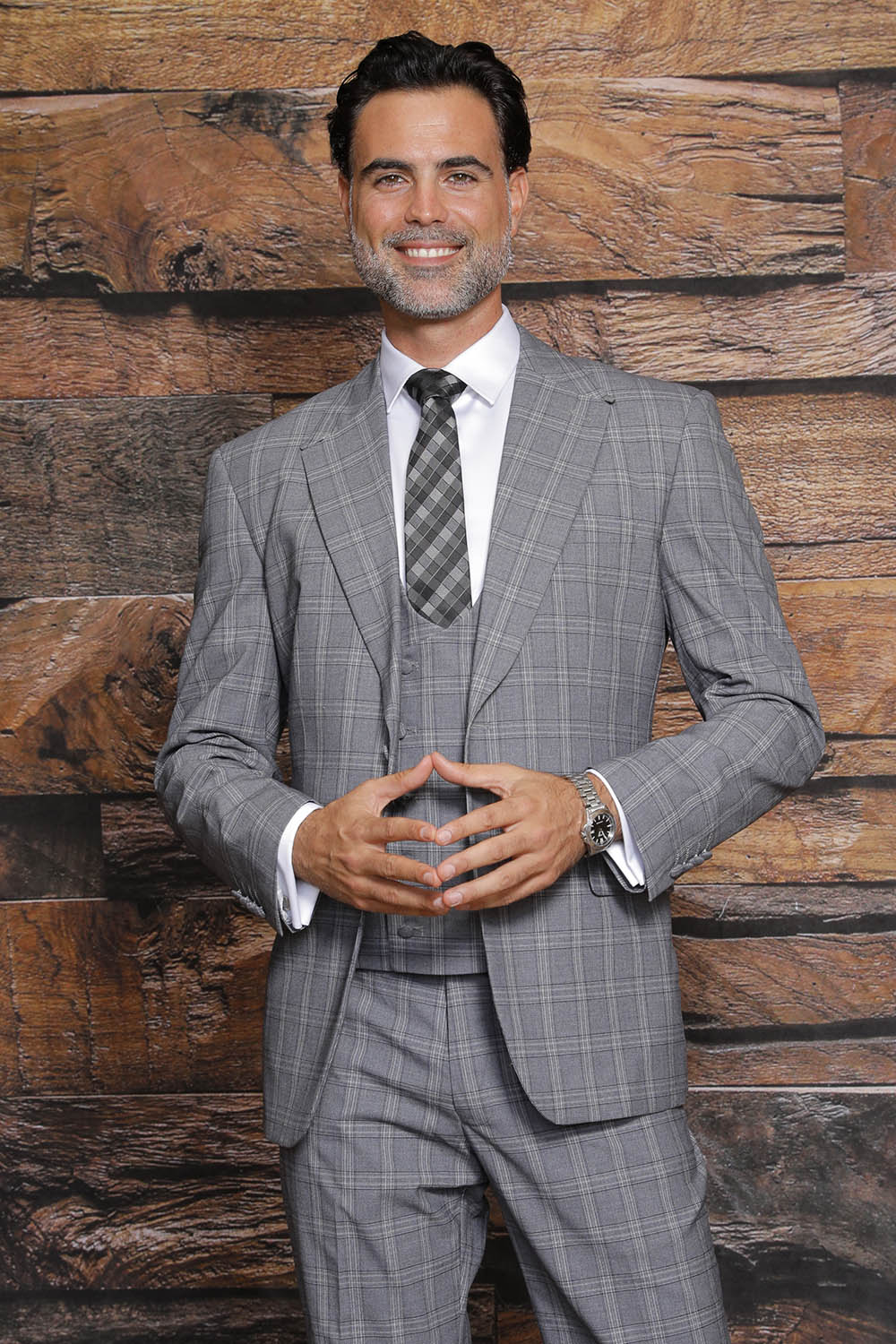 A man in the MANZINI Beverly Charcoal Suit with side vents smiles and clasps his hands, standing in front of a wooden textured background, showcasing the modern fit by Manzini elegantly.