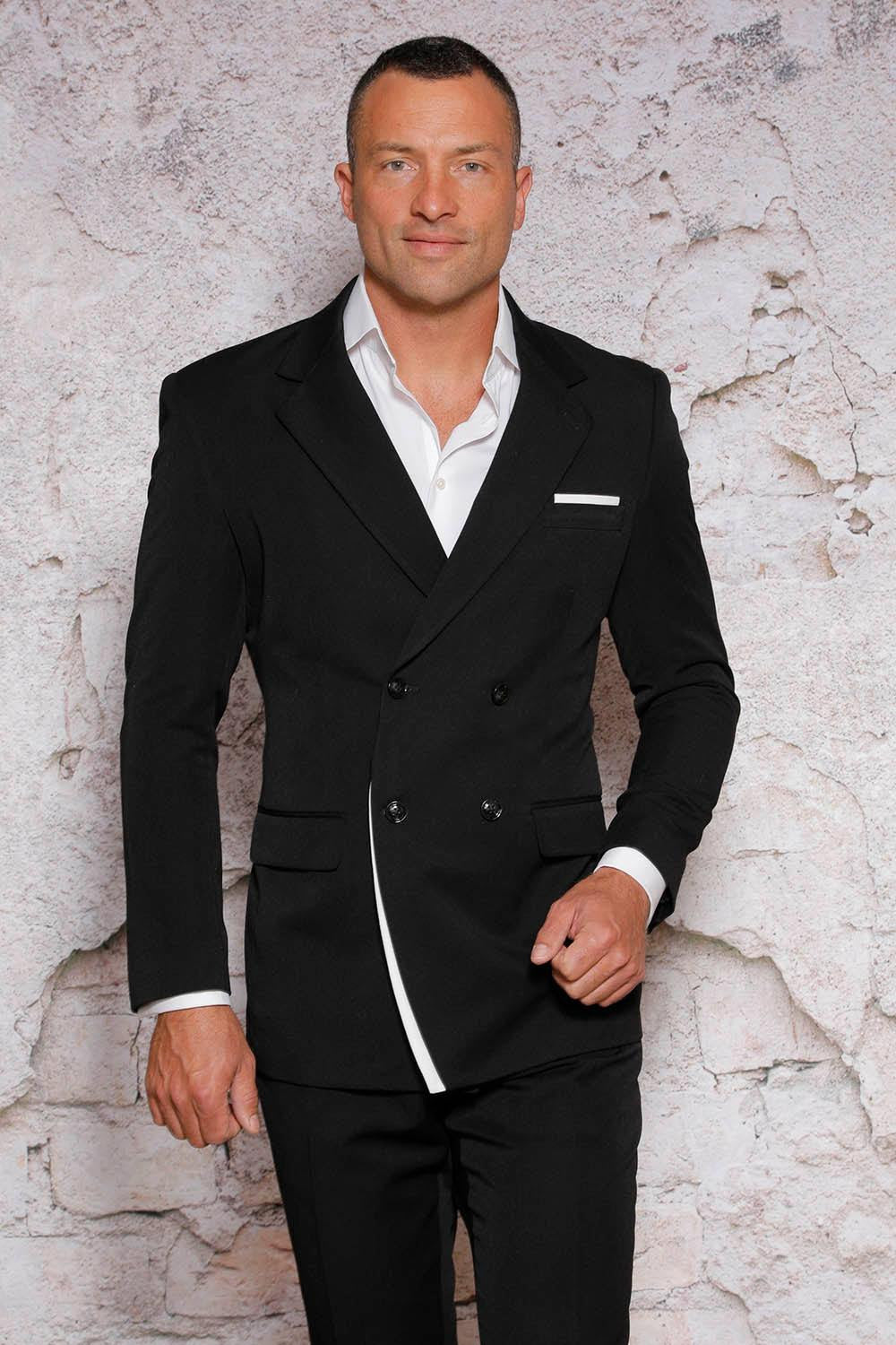 A man wearing a Manzini Fancy Double Breasted Red Label Black/White Suit, featuring a trim notch lapel and paired with a white shirt, stands before a textured stone wall. He gazes straight ahead with a neutral expression.