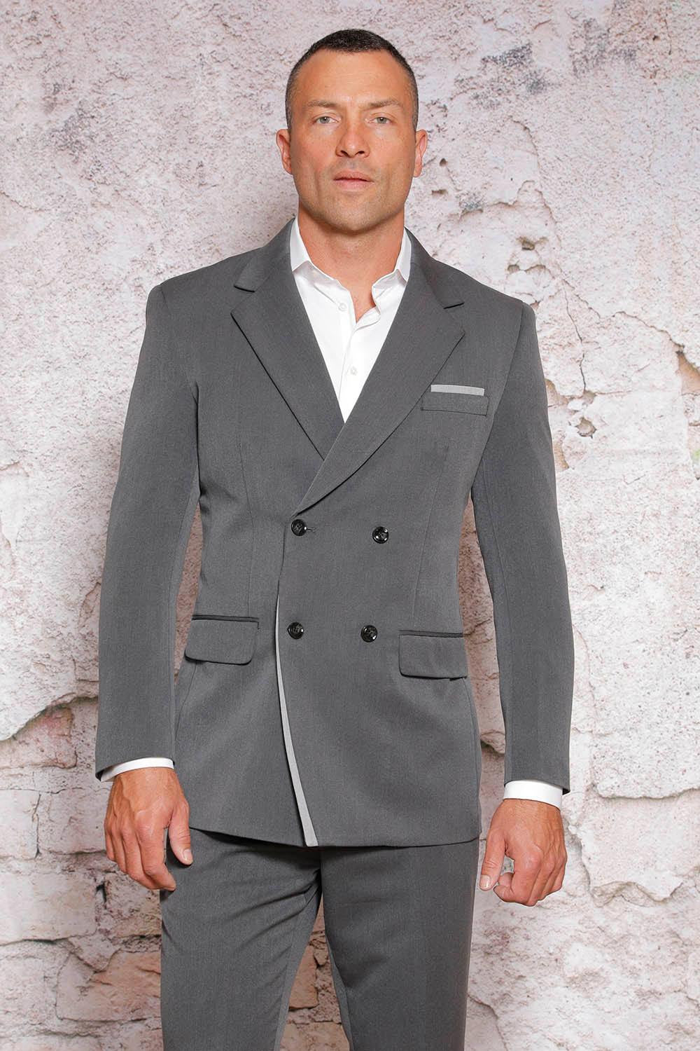 A man wearing a stylish MANZINI Fancy Double Breasted Red Label Charcoal/Ash Suit, featuring trim notch lapels, along with a crisp white shirt, stands against a textured stone wall.