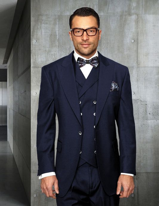 A man wearing the STATEMENT CLOTHING | FLORENCE-NAVY by Statement Clothing, complete with a navy Italian wool three-piece suit and bow tie, stands in a modern, industrial-style hallway.
