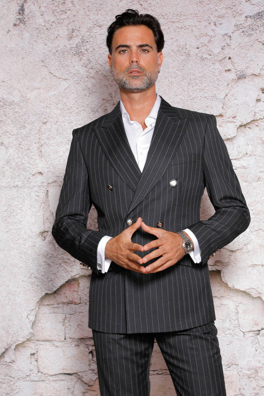 A man with dark hair and a beard wears the MANZINI GQ-100 Black Suit by Manzini, which features a modern fit with flat front pants and side vents, paired with a white shirt. He stands against a textured wall with his hands together.