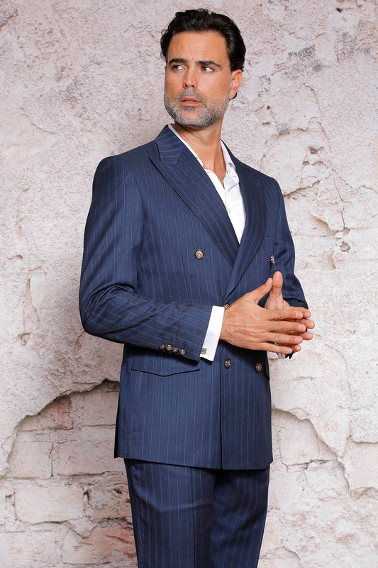 A man wearing the MANZINI GQ-100 Navy Suit, designed with a pinstripe pattern, side vents, and modern fit flat front pants, stands against a textured wall, looking to the side.