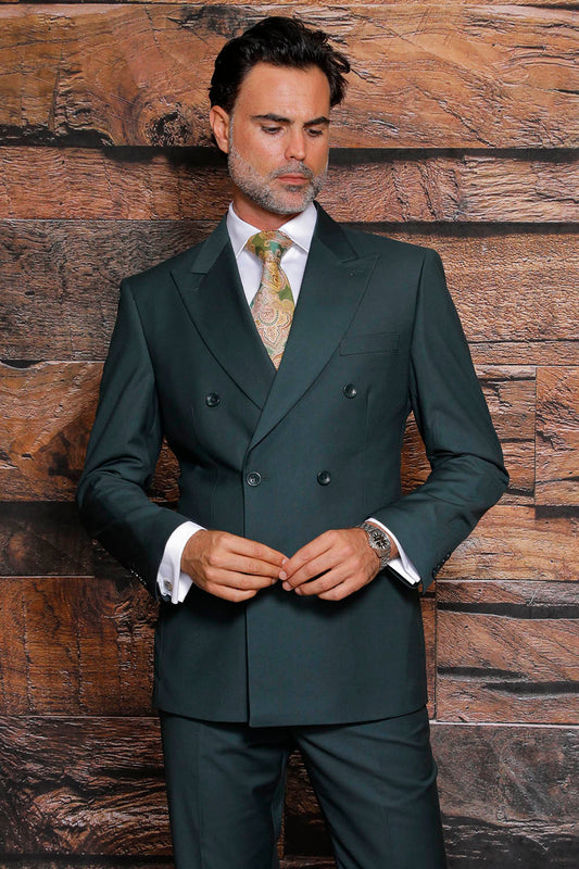 A man wearing a Manzini MANZINI Luna Hunter Suit, featuring a dark green double-breasted design and a modern fit, along with a patterned tie, stands against a wooden background.