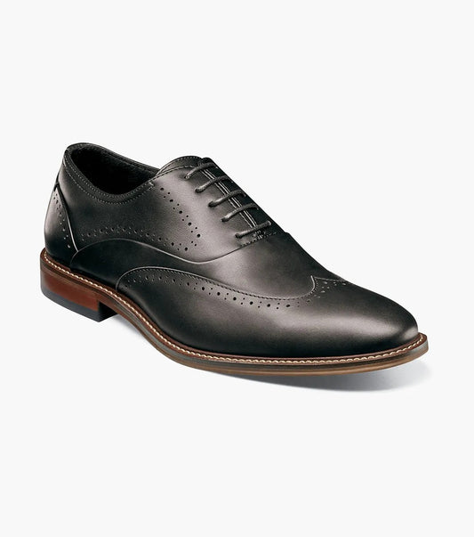 The STACY ADAMS Macarthur Wingtip Oxford (25489-001) is a black burnished leather dress shoe with decorative perforations, brown sole, and black laces, shown against a white background.