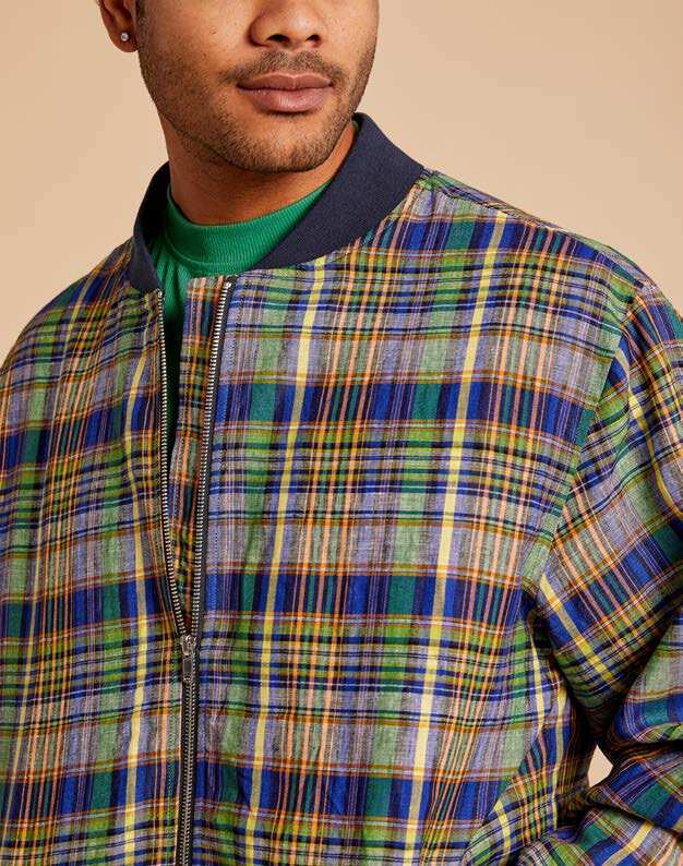 An individual wearing the Inserch Premium Linen Yarn-Dye Madras Plaid Short Zip Jacket in a multi-color pattern over a green shirt is standing against a beige background.