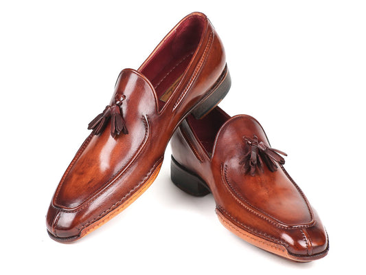 The Paul Parkman Hand-Sewn Tassel Loafers Brown - 082-BRW, expertly crafted from hand-painted leather, exude a sleek, glossy finish. One shoe stands upright as the other leans gracefully against it, accentuating their sophisticated appeal.