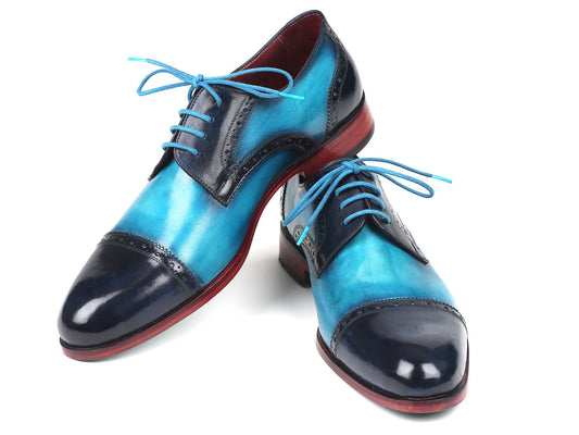 A pair of Paul Parkman Two Tone Cap-Toe Derby Shoes in blue and turquoise, featuring hand-painted leather with blue laces and striking red soles. These expertly crafted men's shoes are arranged with one leaning on the other, making them an ideal addition to elevate any wardrobe.