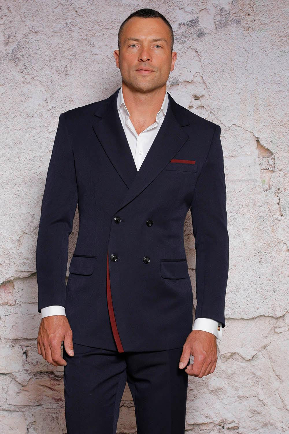 A man in a dark, double-breasted Manzini suit from the MANZINI Fancy Double Breasted Red Label collection is standing against a textured, light-colored wall.