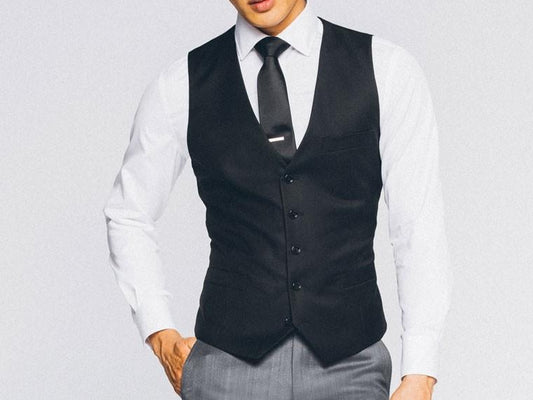A person wearing the Vinci Suits Slim Fit Single Breasted 5 Button Vest (Black) OV-900 over a white shirt with a black tie and gray pants stands against a plain background.